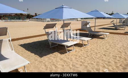 Rangée de chaises longues blanches inoccupées et parasols bleus disposés sur une plage de sable ensoleillée avec une atmosphère côtière calme. Banque D'Images