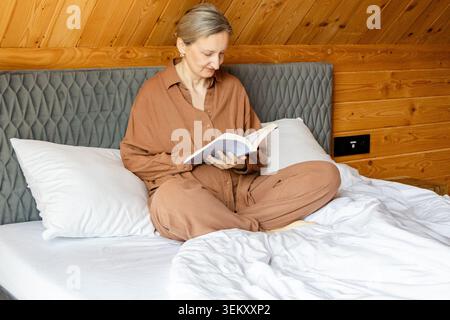Femme d'âge moyen lisant le livre tout en se relaxant sur un lit moelleux dans une cabane en bois grenier. Ambiance matinale confortable, retraite de week-end tranquille, pleine conscience et lent Banque D'Images