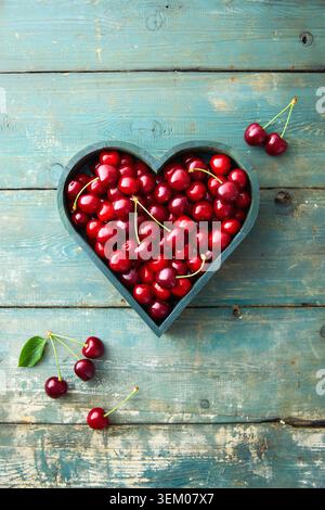 Bol en forme de coeur rempli de cerises rouges fraîches sur fond de bois bleu rustique. Banque D'Images