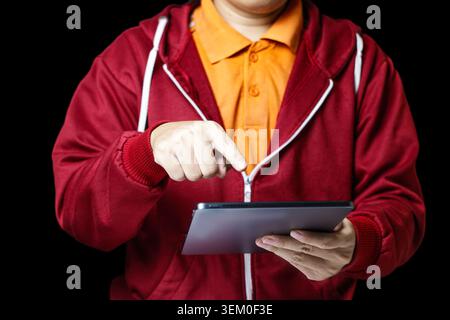 Homme en sweat à capuche rouge tient la tablette. Appuie avec le doigt sur l'écran avec l'intention mise au point. Chemise orange vif contraste le fond sombre. Une tenue élégante suggère un utilisateur moderne et doué de technologie. Idéal pour les affaires, l'éducation. Photo de haute qualité Banque D'Images