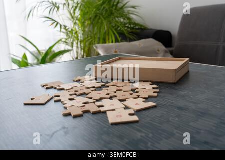 Pièces de puzzle en bois éparpillées avec des symboles géométriques rouges provenant d'une boîte ouverte sur une table sombre avec un fond de plante tropicale et de canapé. Résolution de problème Banque D'Images