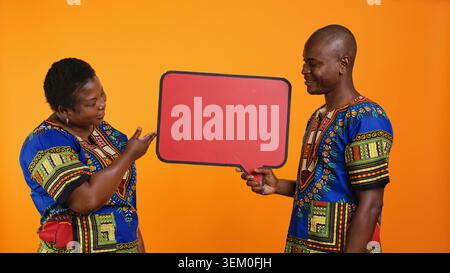 Jeune couple ethnique présentant le panneau de bulles de discours, créant la publicité de marketing et promouvant le panneau d'affichage rouge signe vide dans le studio. Peuple afro-américain pointant vers l'icône en carton. Banque D'Images