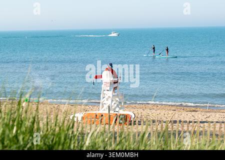 Maître nageur assis sur une grande chaise blanche surplombant une plage de sable aux vagues douces, tandis que les paddleboarders et un bateau à moteur se déplacent sur l'eau turquoise Banque D'Images