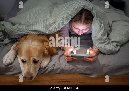Adolescente jouant avec Golden Retriever sous couverture regardant smartphone dans la chambre avant de dormir Banque D'Images