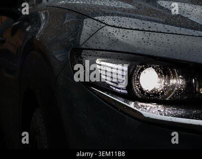 Gros plan sombre d'un phare Mazda sombre recouvert de gouttelettes de pluie par temps humide, avec un style automobile cinématographique par faible luminosité et une faible profondeur O. Banque D'Images