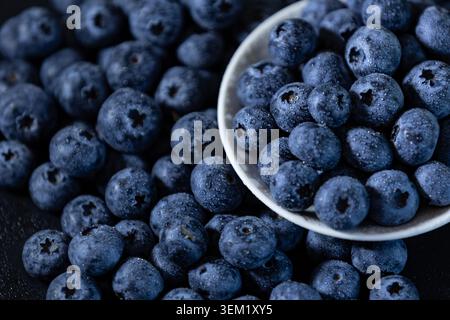 Vue de dessus de bleuets frais mûrs recouverts de gouttelettes d'eau dispersées autour et à l'intérieur d'un bol en céramique blanche sur une surface de table sombre. Banque D'Images