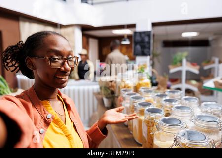 Une jeune femme noire filme des vlogs en utilisant des smartphones pour le marketing et la publicité en ligne. L'image montre une cliente afro-américaine prenant un selfie dans un magasin dynamique et zéro déchet. Banque D'Images