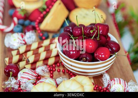 Vue rapprochée de cerises rouges fraîches dans un bol en céramique rayé sur une table à desserts en bois à côté de bâtonnets de gaufrettes au chocolat et de bonbons emballés des fêtes. Banque D'Images