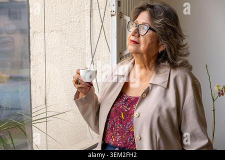Femme senior regardant par la fenêtre, tenant une tasse de café, réfléchissant sur les pensées Banque D'Images