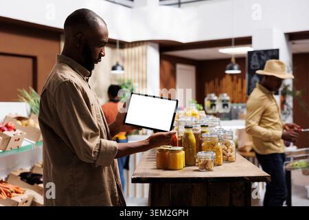 Tout en transportant la tablette avec le modèle chromakey, le gars afro-américain vérifie les articles respectueux de l'environnement, les pots et les aliments sur les étagères. Client masculin saisissant horizontalement un appareil affichant un écran blanc isolé. Banque D'Images