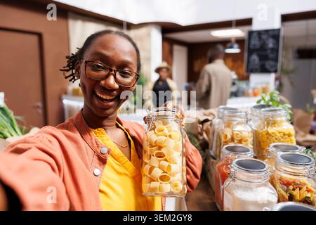 POV d'une femme noire portant des lunettes, tenant un récipient en verre rempli de pâtes dans un supermarché bio-alimentaire. Marketing en ligne, articles organiques, vlogging, appareil photo numérique, tournage client. Banque D'Images