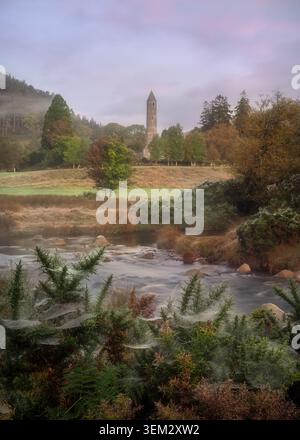 Vue de la tour ronde de Glendalough debout au milieu des arbres d'automne et des collines brumeuses avec une rivière qui coule et des toiles d'araignées au premier plan Glendalough, C. Banque D'Images