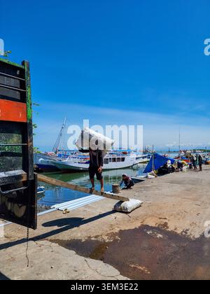 Makassar, Indonésie - 09 février 2024 : Ouvrier portant un sac sur une planche de bois au port de Paotere avec des bateaux traditionnels en arrière-plan. Banque D'Images