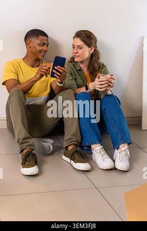 Couple diversifié assis sur le sol carrelé à la maison, partageant smartphone bleu foncé et tasses en céramique Banque D'Images