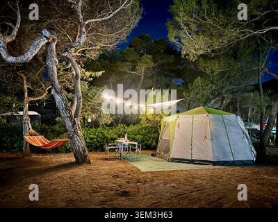 Grande tente de camping illuminée la nuit dans une forêt de pins avec table, hamac et guirlande lumineuse confortable. Vacances d'été en plein air en Toscane, Italie. Banque D'Images