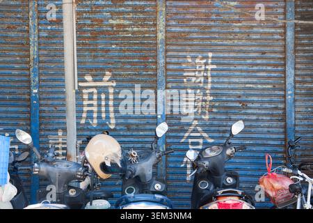 Taipei, Taïwan - 1er juillet 2025 : scooters doublés avant l'obturateur bleu rouillé avec panneau de non-stationnement peint à la main en chinois, scène de rue d'été de Taïwan. Banque D'Images
