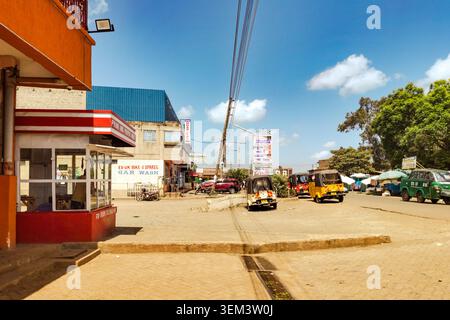 Une scène de rue dans la ville de Kabati, comté de Murang’a, Kenya, mettant en vedette des entreprises locales, un lavage de voiture, garé Tuk-Tuks contre un ciel bleu avec des nuages éparpillés Banque D'Images