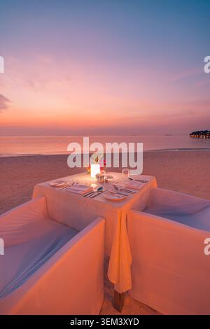 Table intime pour un dîner aux chandelles pour deux sur une plage tropicale lors d'un coucher de soleil romantique. Ciel rose au crépuscule sur l'océan. Voyage de luxe pour lune de miel et escapade Banque D'Images