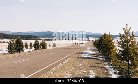 Autoroute asphaltée vide traversant la steppe de Tazheran dans l'oblast d'Irkoutsk, Russie, avec de petits pins, des plaques de neige, des champs ouverts et des collines lointaines sous-jacentes Banque D'Images