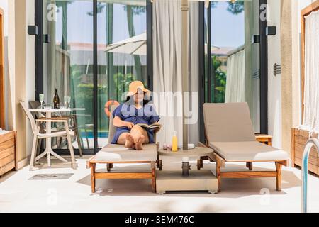 Femme relaxant au bord de la piscine lisant un livre en vacances Banque D'Images