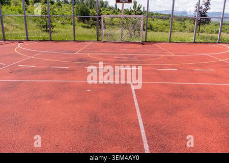 Un encadrement horizontal extérieur d'un terrain de jeu sportif communautaire montrant un sol acrylique rouge altéré avec des marques géométriques de peinture blanche et un soccer Banque D'Images