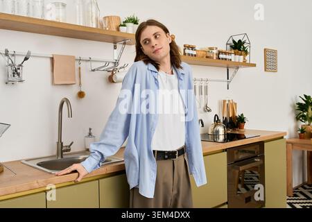 Un jeune homme androgyne se tient confiant dans une cuisine élégante, embrassant son vrai moi. Banque D'Images