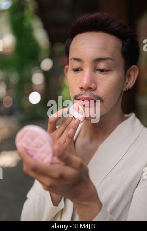 Un portrait haut de gamme et haut de gamme vertical de style de vie d'un homme gay thaïlandais non binaire focalisé appliquant le maquillage à l'extérieur. Le modèle recherche un Banque D'Images