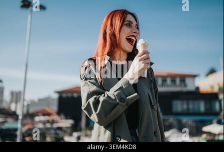 Jeune femme aux cheveux rouges riant en mangeant un cornet de crème glacée à l'extérieur dans un manteau décontracté Banque D'Images