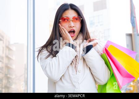 Femme asiatique surprise portant des lunettes de soleil rouges tenant des sacs à provisions colorés dans la ville Banque D'Images