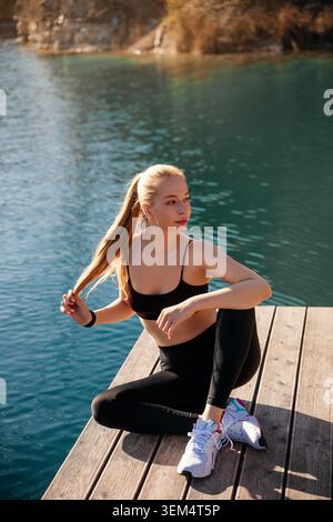 Une femme aux cheveux longs est assise sur un quai en bois près de l'eau tirant ses cheveux en arrière tout en profitant du plein air. Banque D'Images