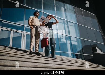Deux collègues debout sur les marches à l'extérieur d'un immeuble de bureaux moderne tenant des documents et des porte-documents Banque D'Images