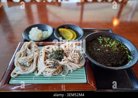 Nouilles de sarrasin froides coréennes sur un plateau en bambou avec sauce salée, radis râpés et wasabi, disposées sur une table en bois brillant. Banque D'Images