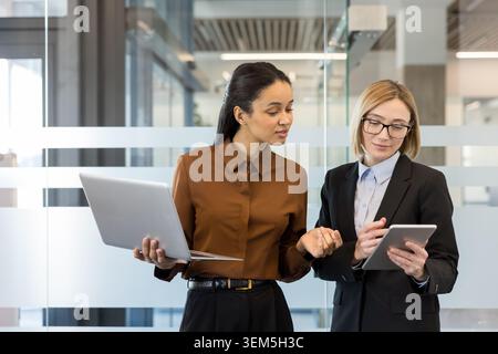 Deux femmes d'affaires diverses debout dans un bureau, discutant de leur travail tandis que l'une tient un ordinateur portable et l'autre utilise une tablette numérique, démontrant le travail d'équipe et la collaboration professionnelle Banque D'Images