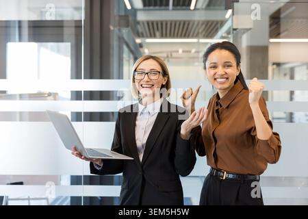 Deux femmes d'affaires diverses dans un bureau moderne célébrant la victoire d'un projet, l'une tenant un ordinateur portable et l'autre pompant ses poings en triomphe, souriant et montrant le travail d'équipe et le succès Banque D'Images