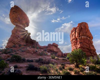 Les rayons du soleil tôt le matin jettent un coup d'œil derrière une colonne de grès rouge avec un rocher équilibré au sommet dans le parc national des Arches, Moab, Utah Banque D'Images