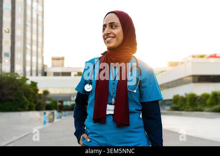 Professionnelle de santé musulmane portant un hijab et des gommages sarcelle, regardant avec confiance sur le côté à l'extérieur d'un établissement médical, incarnant la dédicace Banque D'Images
