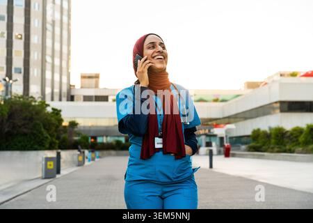 Professionnelle médicale arabe souriante tout en ayant une conversation téléphonique, portant des gommages et du hijab, marchant en toute confiance devant un hospita moderne Banque D'Images