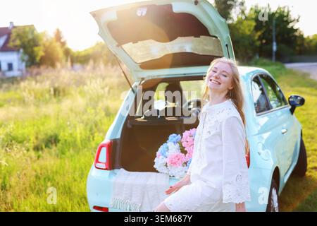 Voyageur élégant appréciant un pique-nique paisible avec du vin et des fleurs d'un coffre de voiture ouvert dans un paysage rural pittoresque à l'heure dorée Banque D'Images