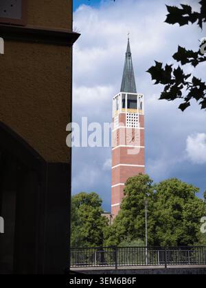 Pforzheim, Allemagne -Juni 6, 2026 Une grande tour d'horloge en brique rouge avec une flèche sombre mince s'élève au-dessus des arbres verts feuillus, encadrée à gauche par la corne Banque D'Images