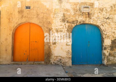 Vue de deux portes en bois cintrées, une orange et une bleue, situées dans un mur de calcaire altéré à Zebbug, région de Gozo, Malte. Banque D'Images