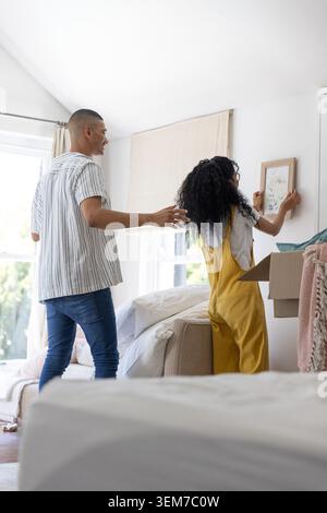 Couple accrochant imprimé floral encadré dans la chambre à coucher par la fenêtre, femme en salopette moutarde, homme faisant des gestes Banque D'Images
