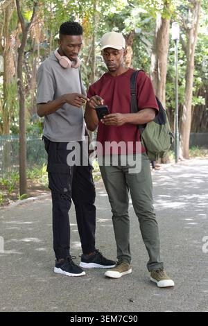 Deux hommes africains debout sur la passerelle du parc pointant vers le smartphone, portant un sac à dos et des écouteurs Banque D'Images