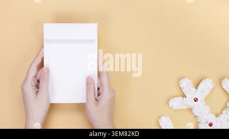 Gros plan des mains tenant une enveloppe blanche vierge sur un fond beige avec une décoration de Noël blanche moelleuse. Ceci est une excellente maquette pour mettre votre texte Banque D'Images
