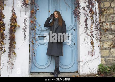 Debout en tenue d'hiver, une femme pose près d'une porte bleue frappante avec du lierre séché à Montmartre, Paris. La scène combine la beauté rustique avec la f parisienne Banque D'Images