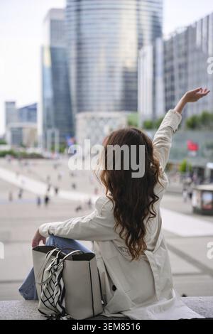 Une femme lève le bras en étant assise dans le quartier des affaires de la Défense à Paris face à la skyline et profitant de l'atmosphère de la ville Banque D'Images