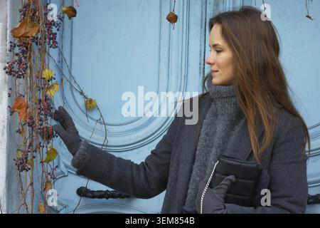 Dans une tenue d'hiver confortable, une femme pose avec confiance contre une porte bleue vibrante avec lierre en cascade à Montmartre, Paris, créant une scène élégante riche wi Banque D'Images