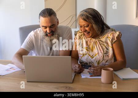 Homme adulte mature et femme senior assis à la maison, examinant ordinateur portable et carte de crédit sur la table Banque D'Images
