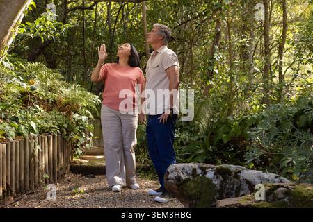 Divers couples âgés marchant chemin de gravier dans le jardin ombragé près de poteaux en bois, la femme pointant vers le haut Banque D'Images