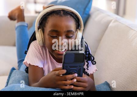Enfant afro-américain allongé sur un canapé à la maison portant des écouteurs supérieurs roses tenant l'étui de smartphone Banque D'Images
