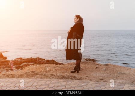 Femme côte coucher de soleil debout sur une rive rocheuse dans un manteau noir regardant l'eau calme de l'océan pendant l'heure dorée Banque D'Images